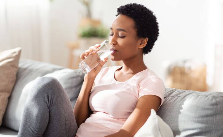 A relaxed Afro-american woman sitting on a sofa and drinking water from a glass.