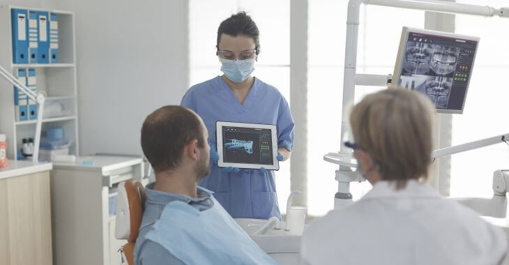 A dentist holding teeth x-rays and explaining the issue and treatment to a male patient sitting in a dental chair