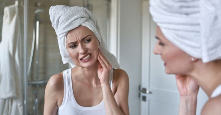 A woman feeling dental pain looking at her teeth in a bathroom mirror.